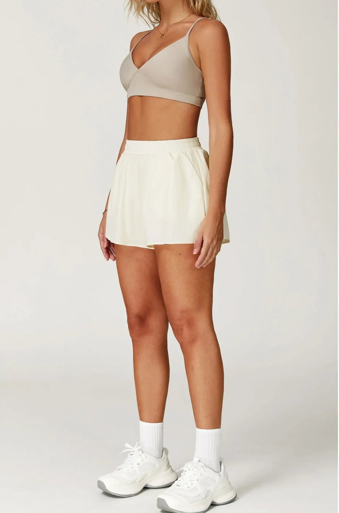 Person wearing a beige sports bra and shorts with white sneakers on a light gray background