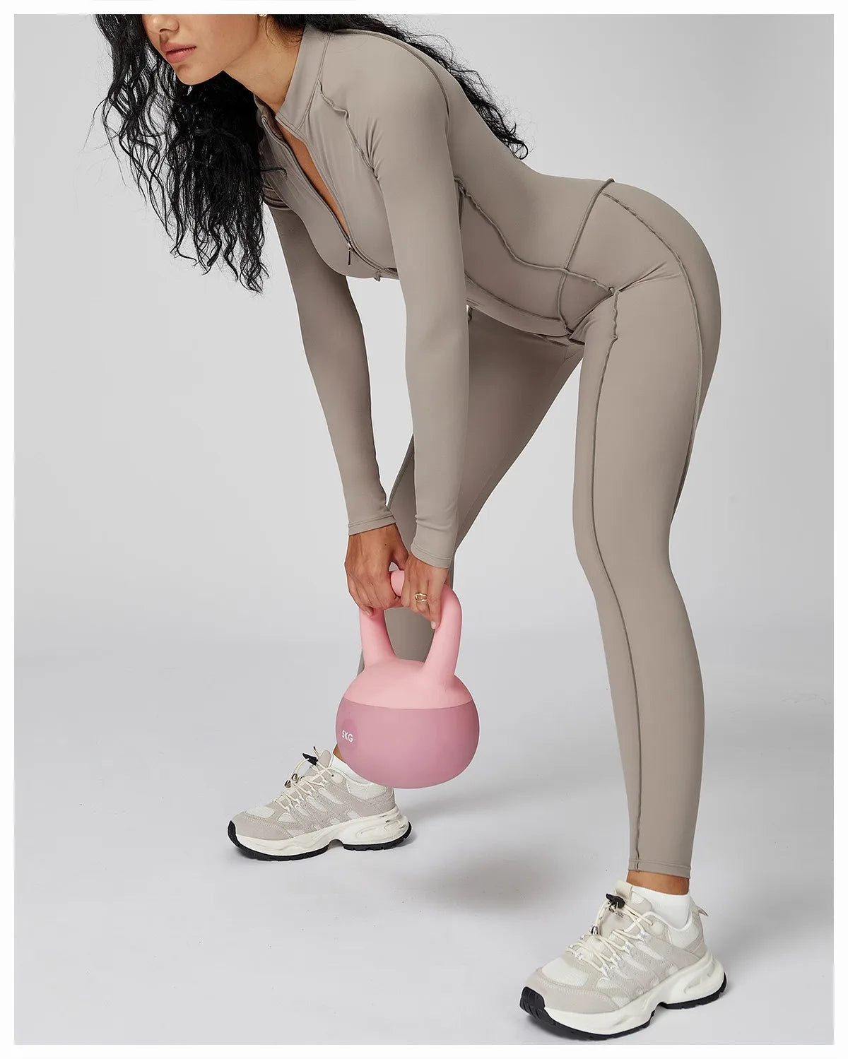 Person lifting a pink kettlebell wearing a beige athletic jumpsuit on a light gray background