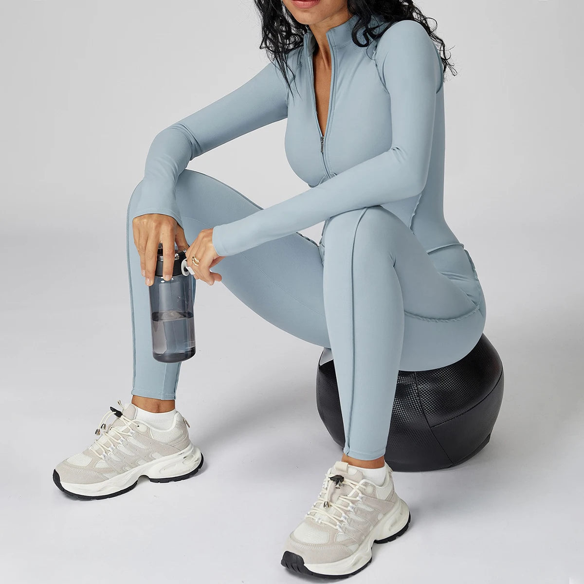 Person in a light blue athletic jumpsuit sitting on a black exercise ball with a white background