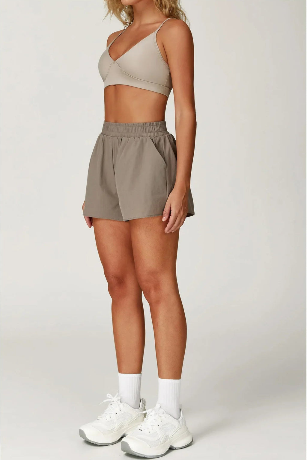 Person wearing a beige sports bra and shorts with white sneakers on a light gray background